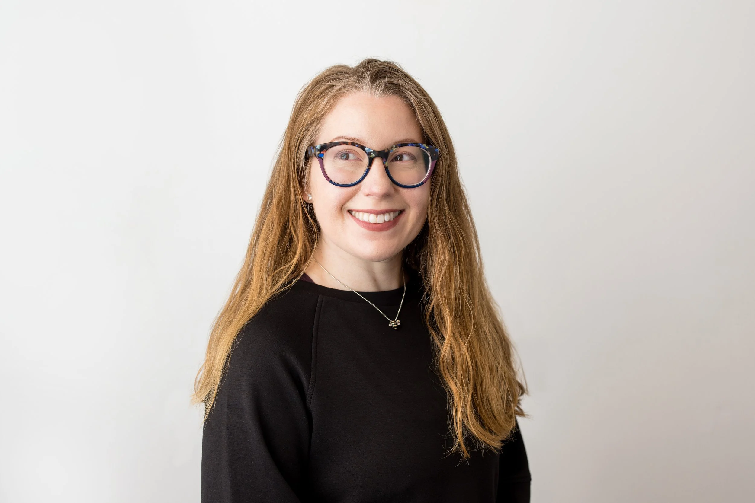 A woman with long, wavy, red hair and glasses  smiling in a gray long-sleeve shirt against a plain white background.