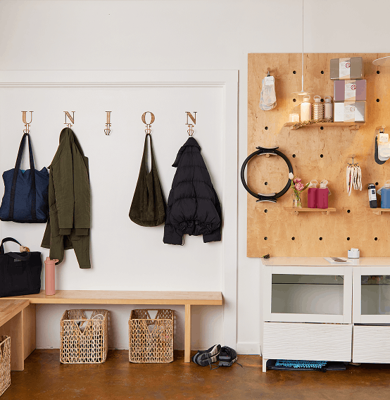 Entryway with hooks holding a blue bag, green jacket, black bag, and black puffy coat, a wooden bench below with two wicker baskets, and a pegboard on the right with shelves and hooks for various items.