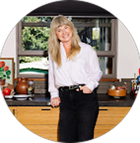 Woman with blonde hair wearing a white shirt and black pants standing in a kitchen with a window view of greenery.