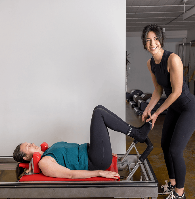 Woman exercising on a Pilates reformer machine while smiling at the camera, in a gym setting.