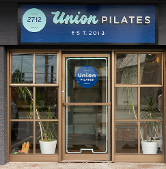 Front view of a Pilates studio called Union Pilates, established in 2013, with a glass door and large windows, potted plants on each side of the entrance.
