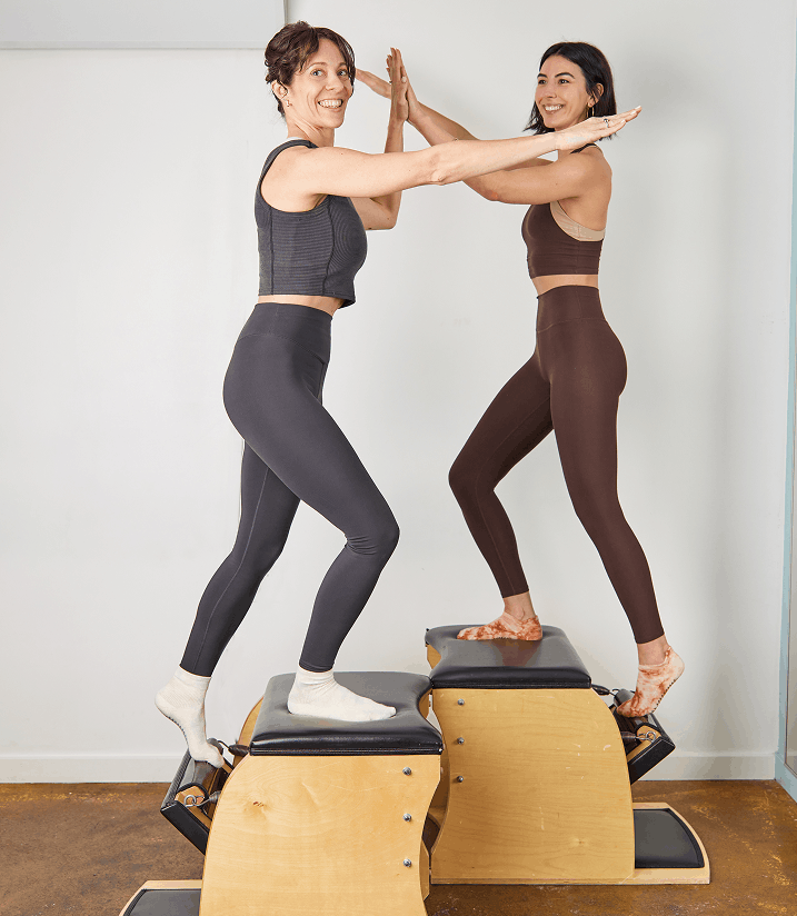 Two women in workout clothes doing a high five on a Pilates reformer machine.