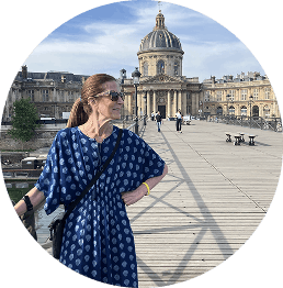 Woman in a blue patterned dress standing on a rooftop with historic buildings and a domed structure in the background under a partly cloudy sky.