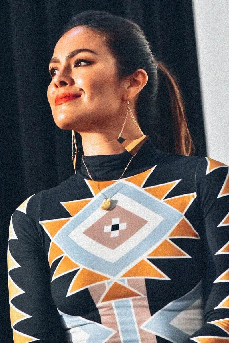 A woman with dark hair tied in a ponytail, wearing geometric earrings, and a patterned top with bold geometric shapes in black, orange, white, and beige.
