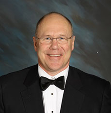 A man in a tuxedo and bow tie, smiling, with glasses and short hair, posed against a dark, abstract background.