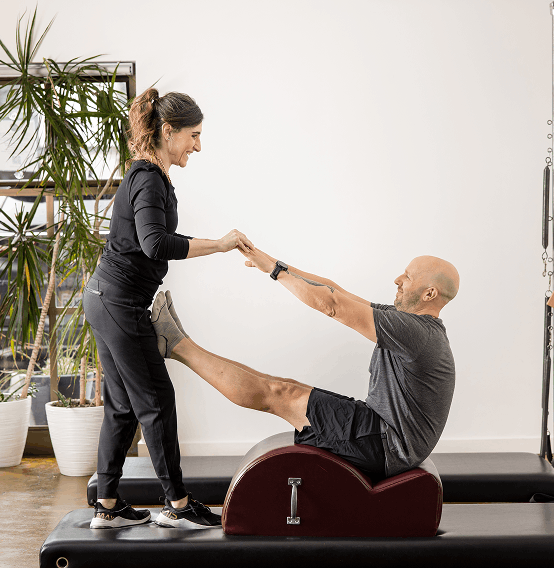 A woman assisting a man during physical therapy exercises in a gym, with the man holding her hands and seated on a specialized chair.