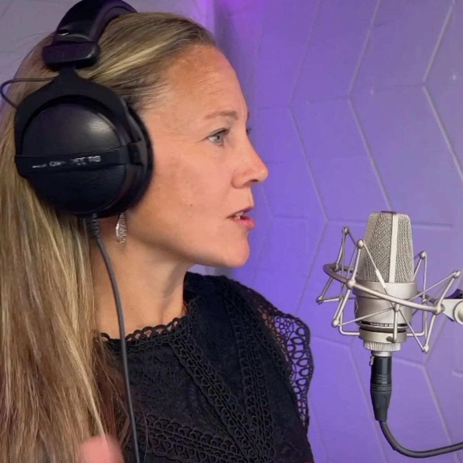 A woman with long blonde hair wearing headphones and singing into a studio microphone.