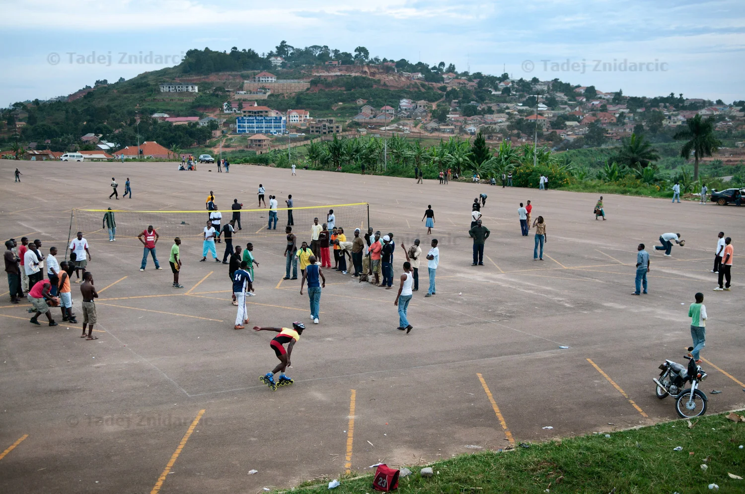 Inline speed skater passes behind a group playing volleyball at Mandela National Stadium's parking lot in Kampala, Uganda. When there is no event at the stadium, the parking lot is open to the public. Skaters share it with others who come to the parking lot for recreation or are just passing through. They have to weave around pedestrians, avoid bicycles and duck stray balls.