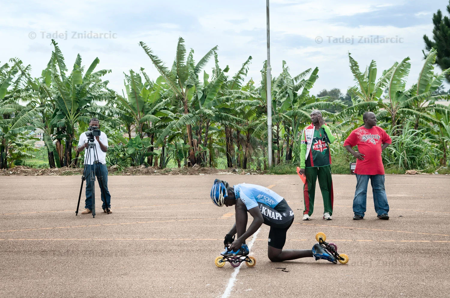 Cameraman from a local TV films Mugabi Julius as he is preparing his skates for a race during 2nd East African inline speed skating competition at Mandela National Stadium's parking lot in Kampala, Uganda, while Ugandan coach Ddungu Moses (in red) and Kenyan coach wait for the start of the race. Though inline speed skating is not a mainstream sport in Uganda, local media houses have started to take notice.