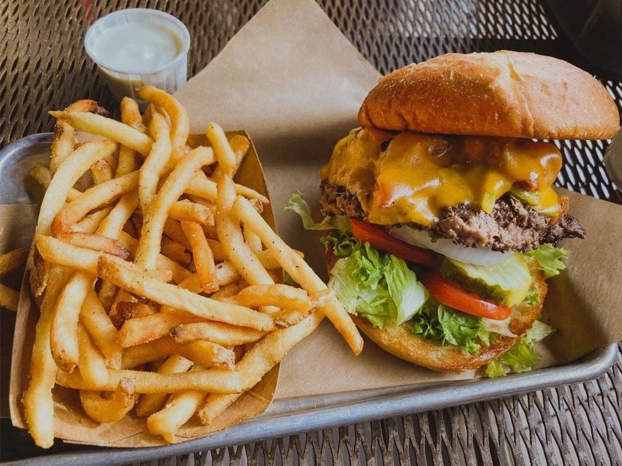 A grass-fed beef burger with lettuce, tomato, pickles, onions, cheddar cheese, and a beef patty on a bun, served with a side of French fries and a small cup of dipping sauce.