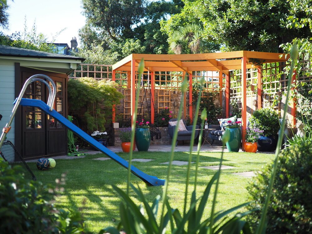  A children’s slide sits on a well maintained lawn with stepping stones to a shed and a patio dining area with a pergola overhead. 