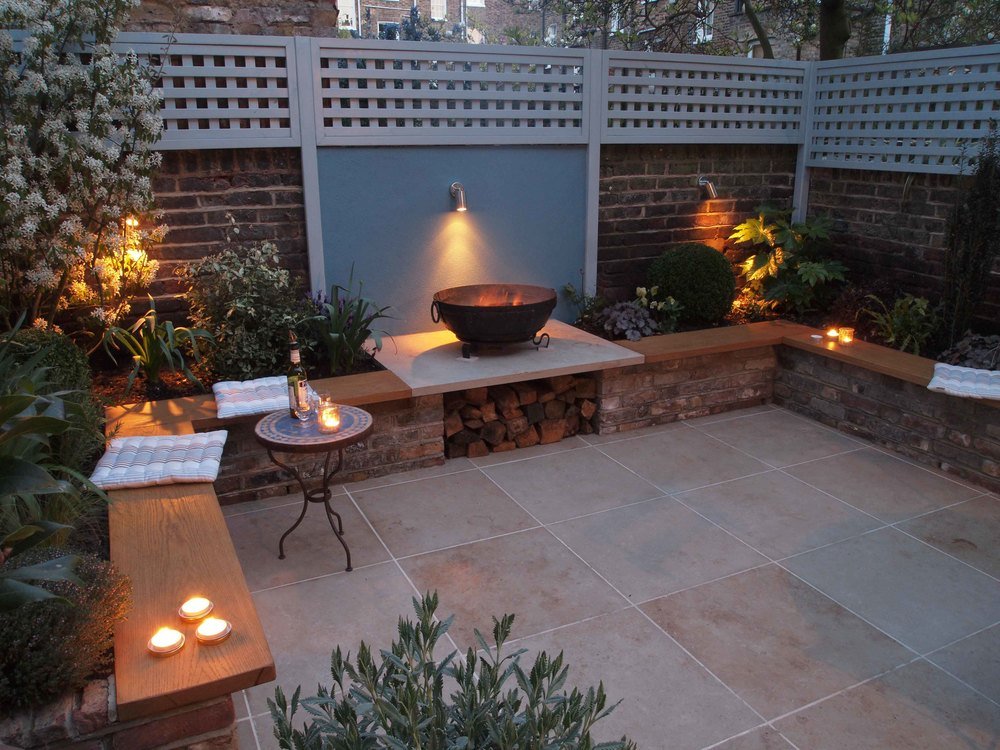  Stunning small garden design in North London with outdoor lighting on the boundary wall highlighting the fire bowl, limestone patio and reclaimed brick planters filled with plants from Provence. 