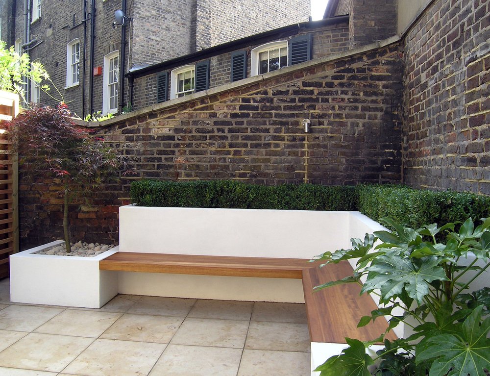  Close up of the limestone patio in this family friendly garden with a modern floating, L-shaped underlit Iroko bench surrounded by white rendered planters. 