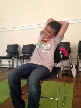 How to : While sitting in your seat, uncross your knees and ankles and sit up tall. Bring your right arm up beside your head and bend your elbow so that your right hand is reaching down the center of your back. Use your left hand to gently hold the right elbow. Inhale, sit tall . Exhale, and stretch over to the left, lengthening your right side body and stretching your tricep(under part of upper arm). Hold and breath for 3-5 breaths. Repeat 2-4 times on both sides. Kids can make Sssshhhhhhh sounds when stretching their side body or horse breath(relax the lips and blow breath out creating a vibration through the lips, and a horse neigh sound). Benefits: Elongates the spine and gives your lungs and other organs space. 