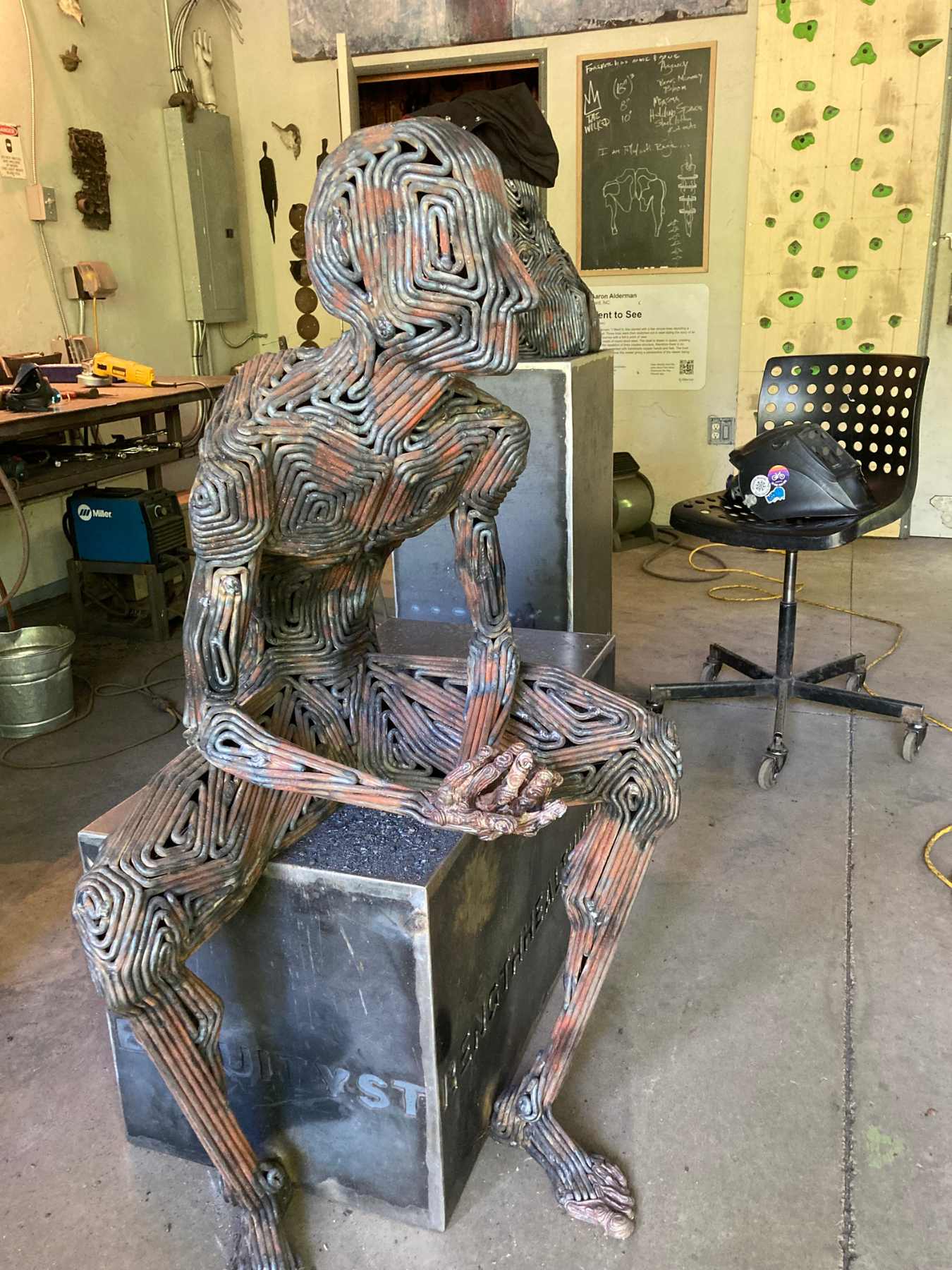 Steel sculpture of seated man leaning on his elbows on his knees in the studio of sculptor J. Aaron Alderman. Viewed from the front.
