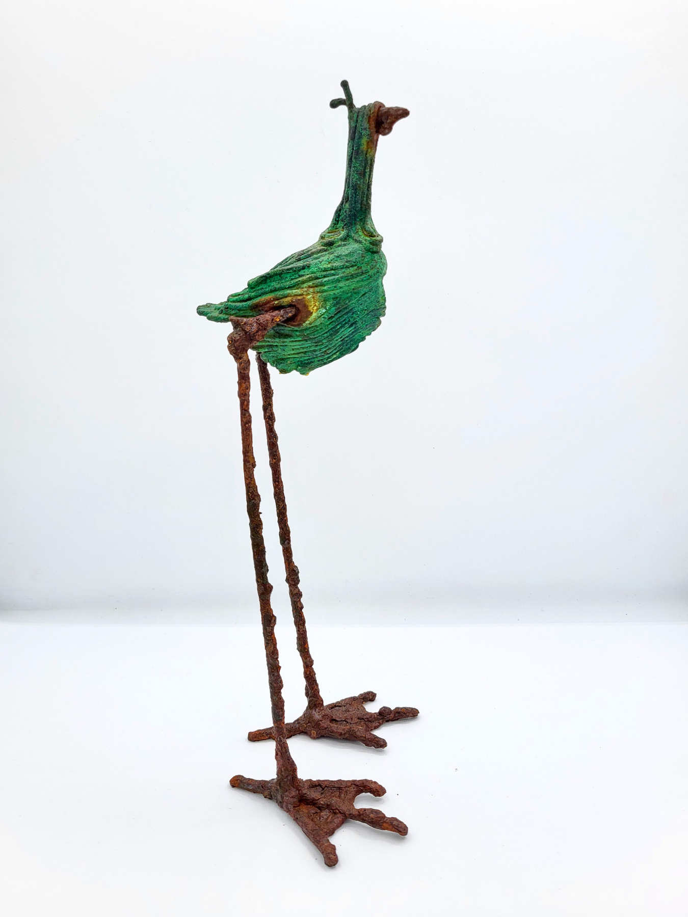 A long-legged bird statue made from steel with a copper patina faces right against a white gradient background.