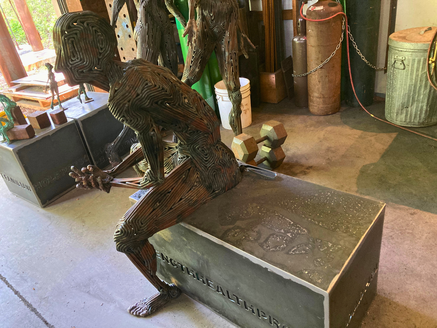 Steel sculpture of seated man leaning on his elbows on his knees in the studio of sculptor J. Aaron Alderman. Viewed from the side.