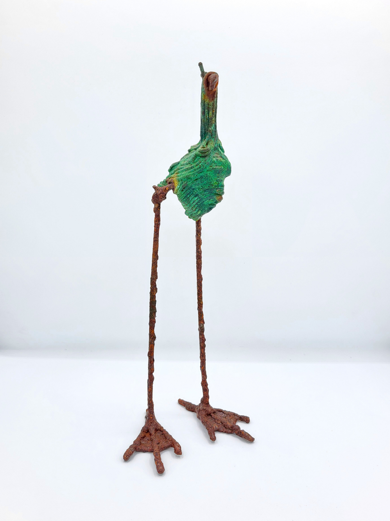 A long-legged bird statue made from steel with a copper patina faces forward against a white gradient background.
