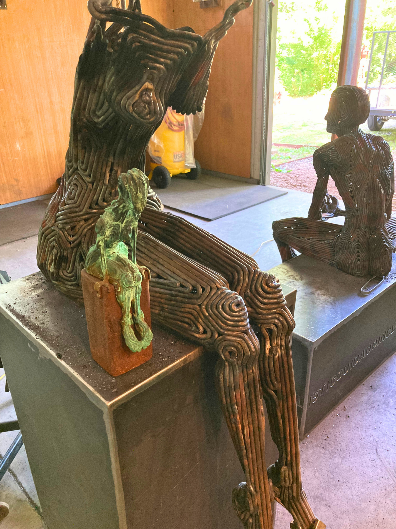 Side view of a steel sculpture and maquette of a seated woman with legs dangling in the studio of sculptor J. Aaron Alderman. Viewed from front.