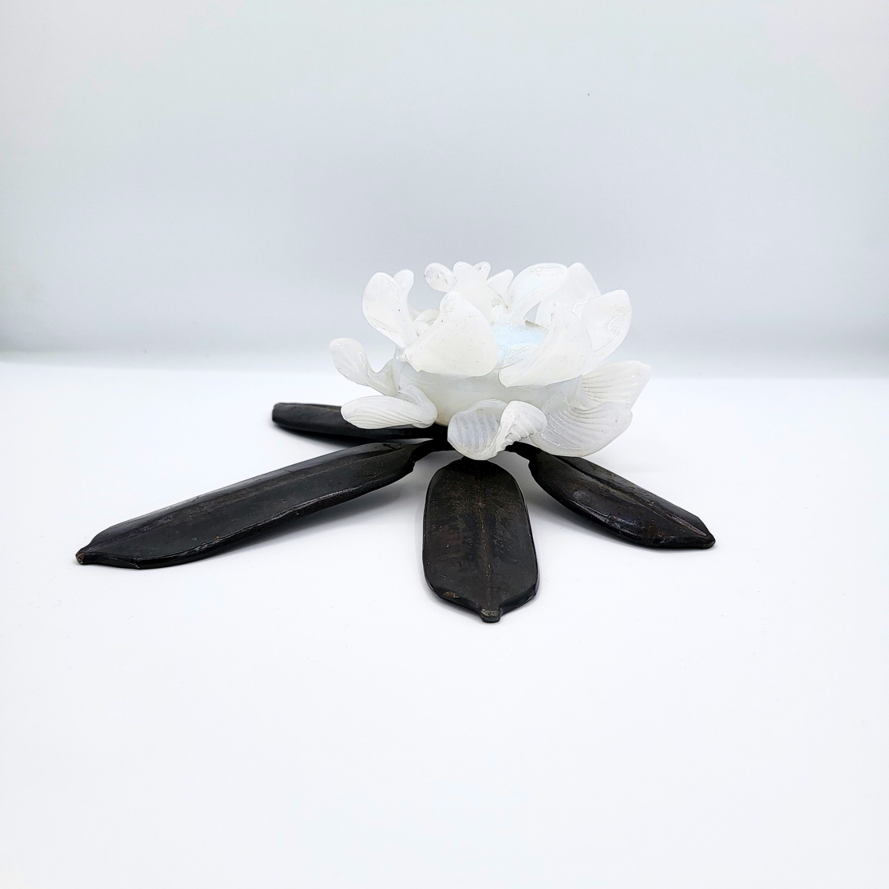 Hand-forged rhododendron leaf with white glass flower nestled in the center against a white background.