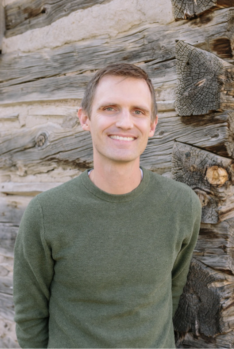 Matt Meyer with green sweater on posing in front of log cabin