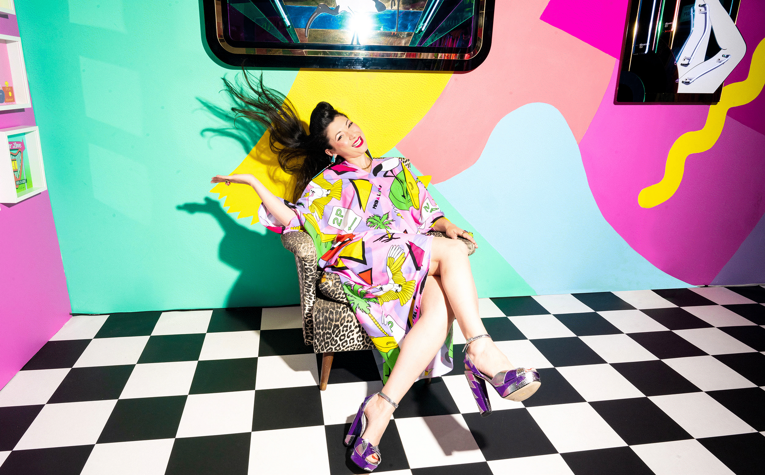 A animated GIF of an artist sitting in her bright maximalist 1980s-inspired studio wearing a colourful Pop Art kaftan flicking her hair and introducing her new book Australianarama A Campendium of Kitsch, releasing in September 2026