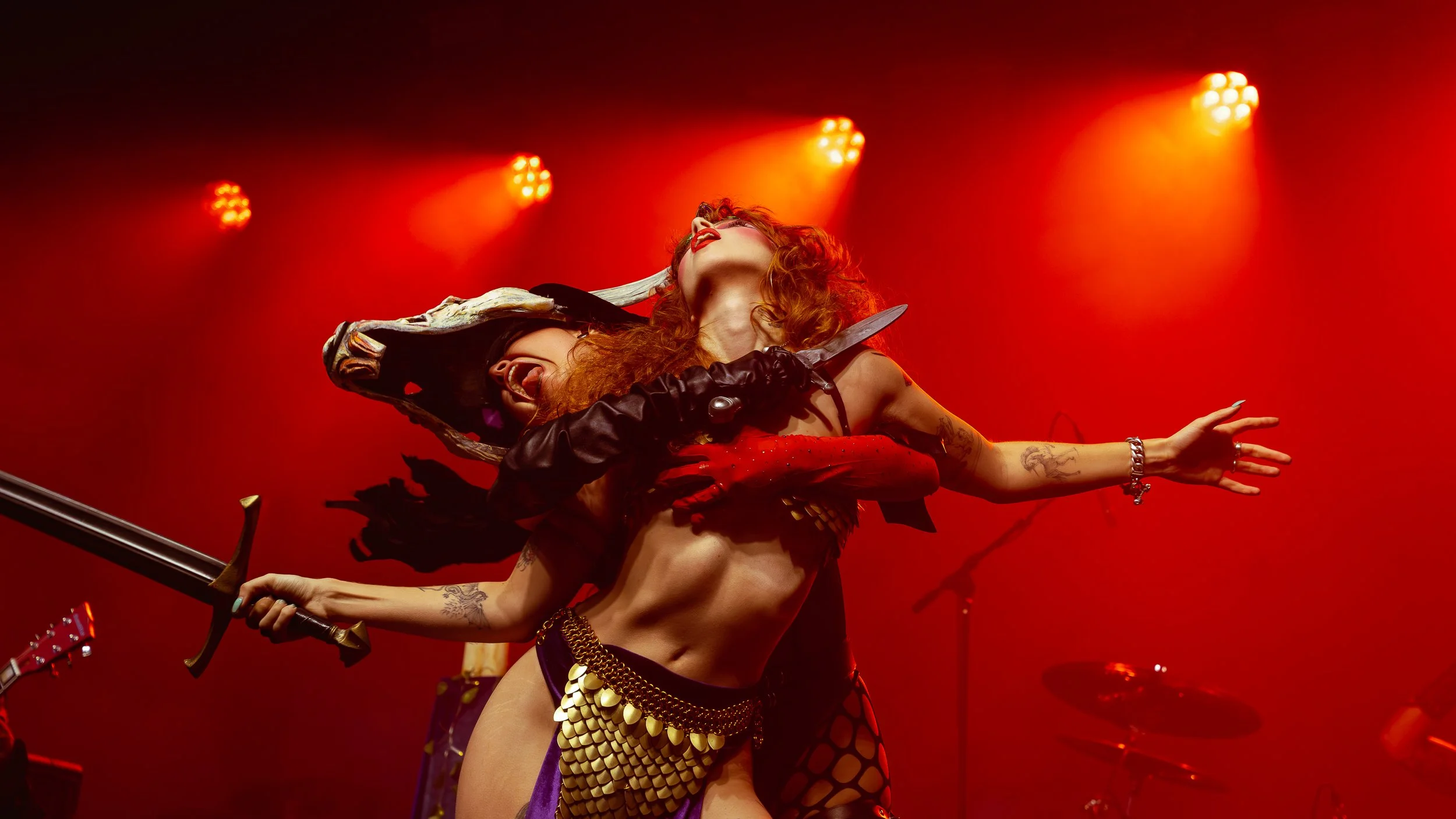 A performer on stage with red lighting, holding a sword, dressed in a costume with gold accents, and red curly hair, striking a dramatic pose.