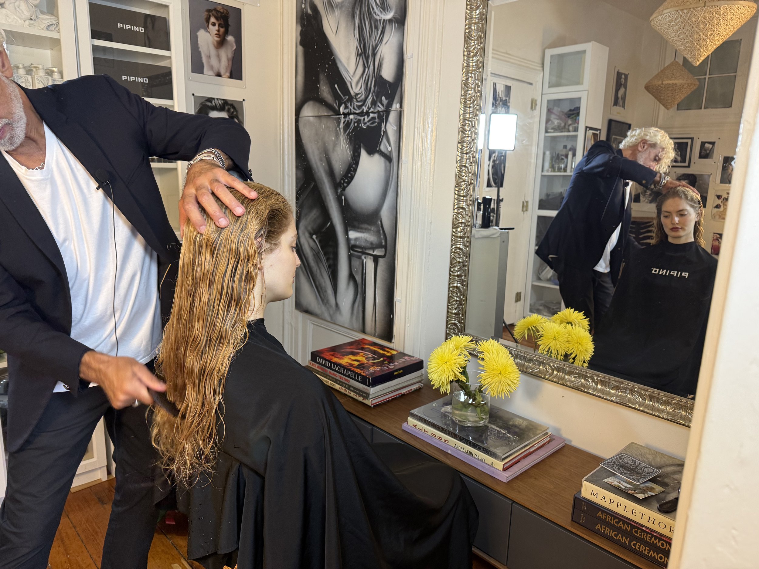 A hairstylist combing a woman's wet hair in a chic salon. The woman, draped in a black cape, watches her reflection in a large ornate mirror, surrounded by books and fresh yellow flowers.