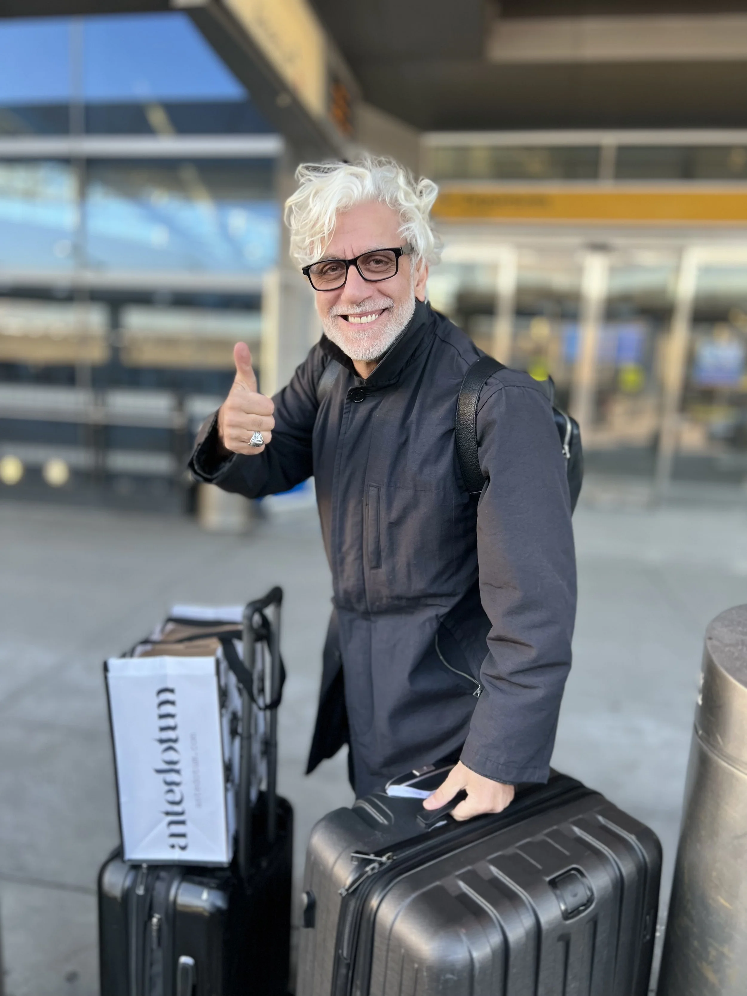 A stylish man with white hair and glasses, wearing a dark jacket, giving a thumbs-up while standing beside suitcases and a shopping bag outside a building.