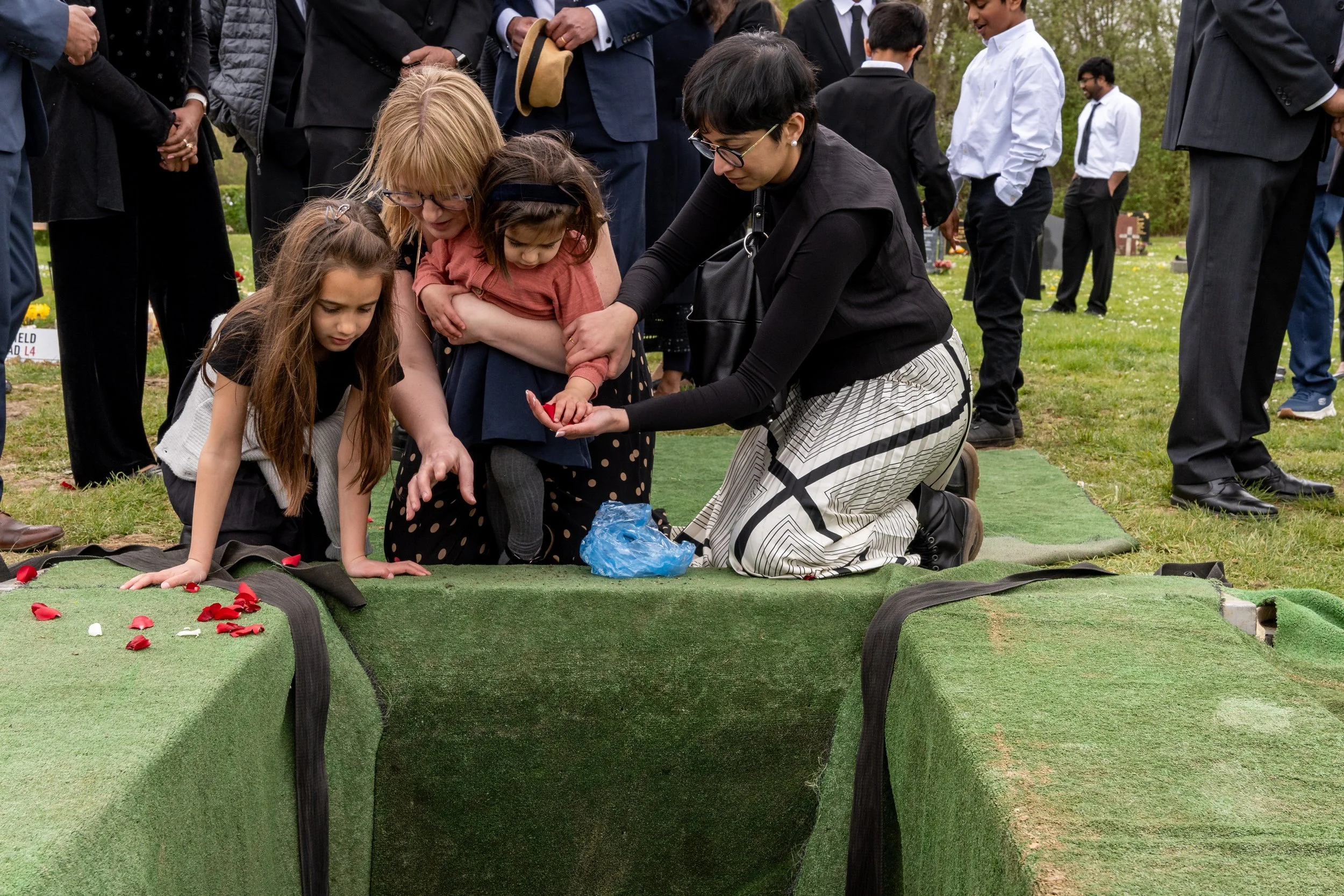 Women and young children kneel at the graveside, gently placing rose petals into the grave, documentary funeral photography by Paul Hands