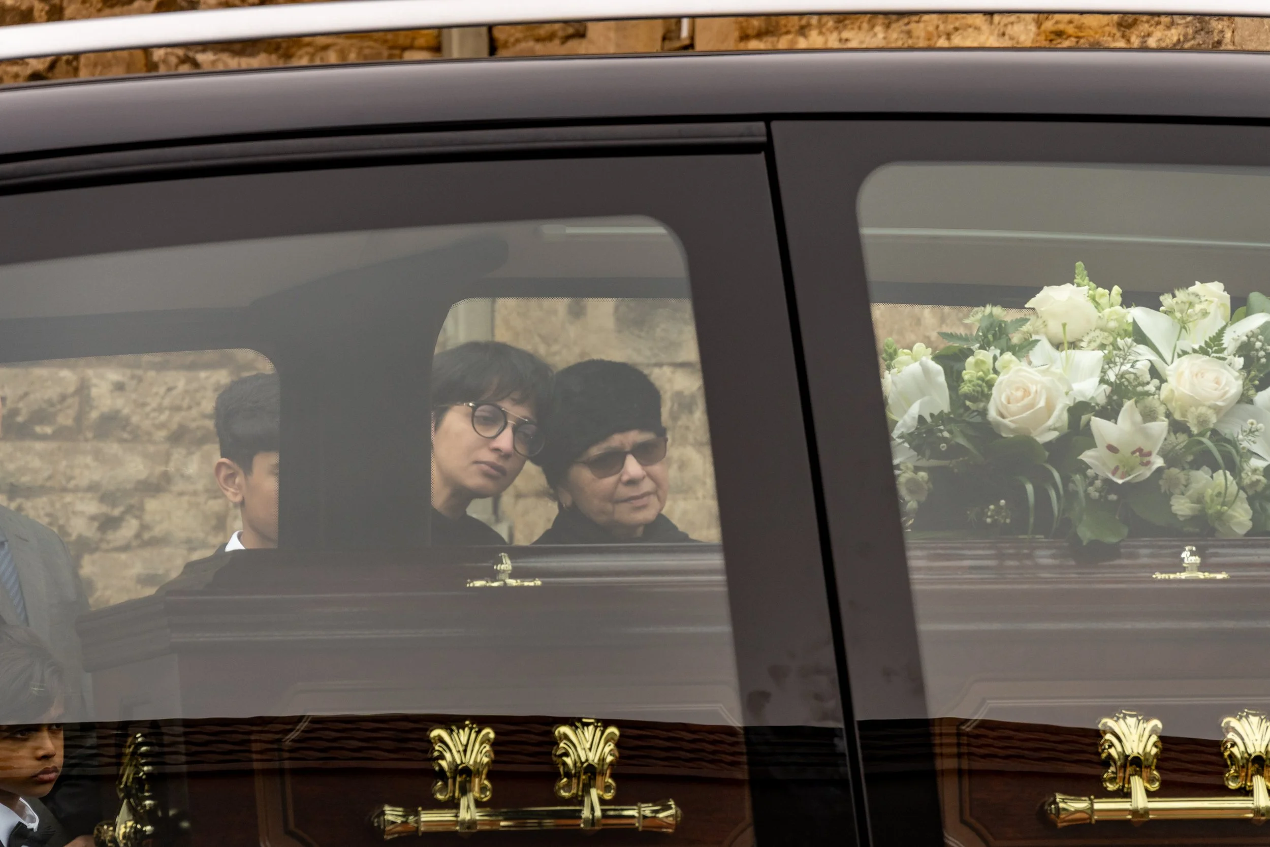 Family members seated beside the coffin inside a funeral car, white lilies resting against the window, documentary funeral photography by Paul Hands