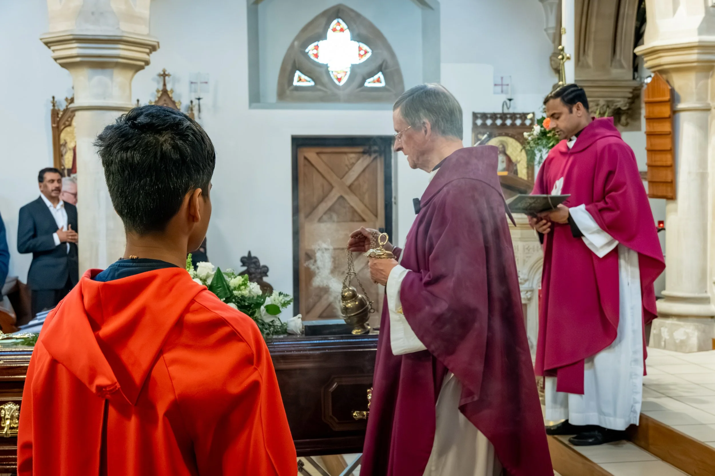 A priest in magenta robes conducts a blessing at the altar during a multi-faith funeral ceremony in Bedford, candid funeral photography by Paul Hands