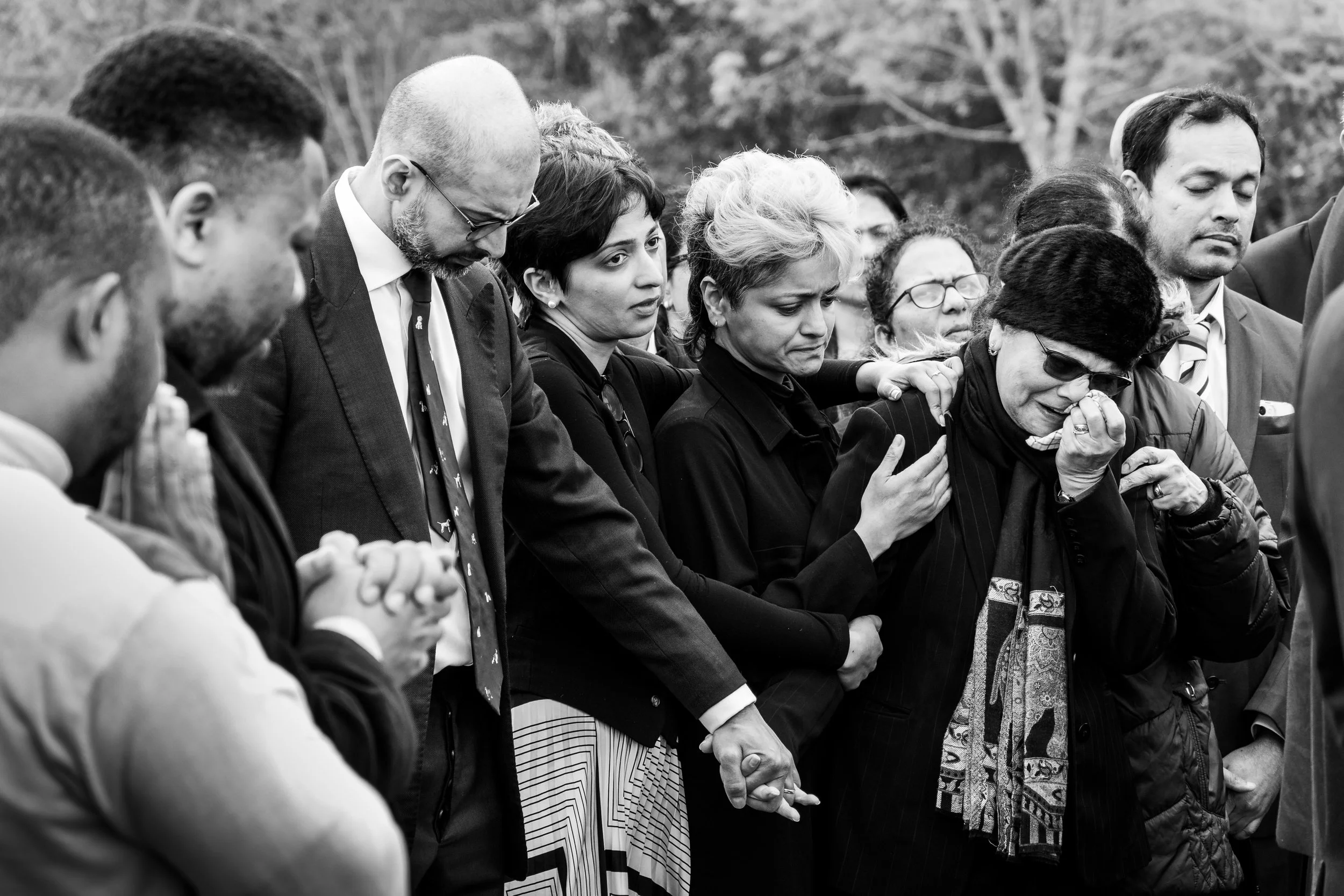 A close group of mourners sharing grief at a graveside, hands clasped together, one woman weeping, candid black and white funeral photography by Paul Hands