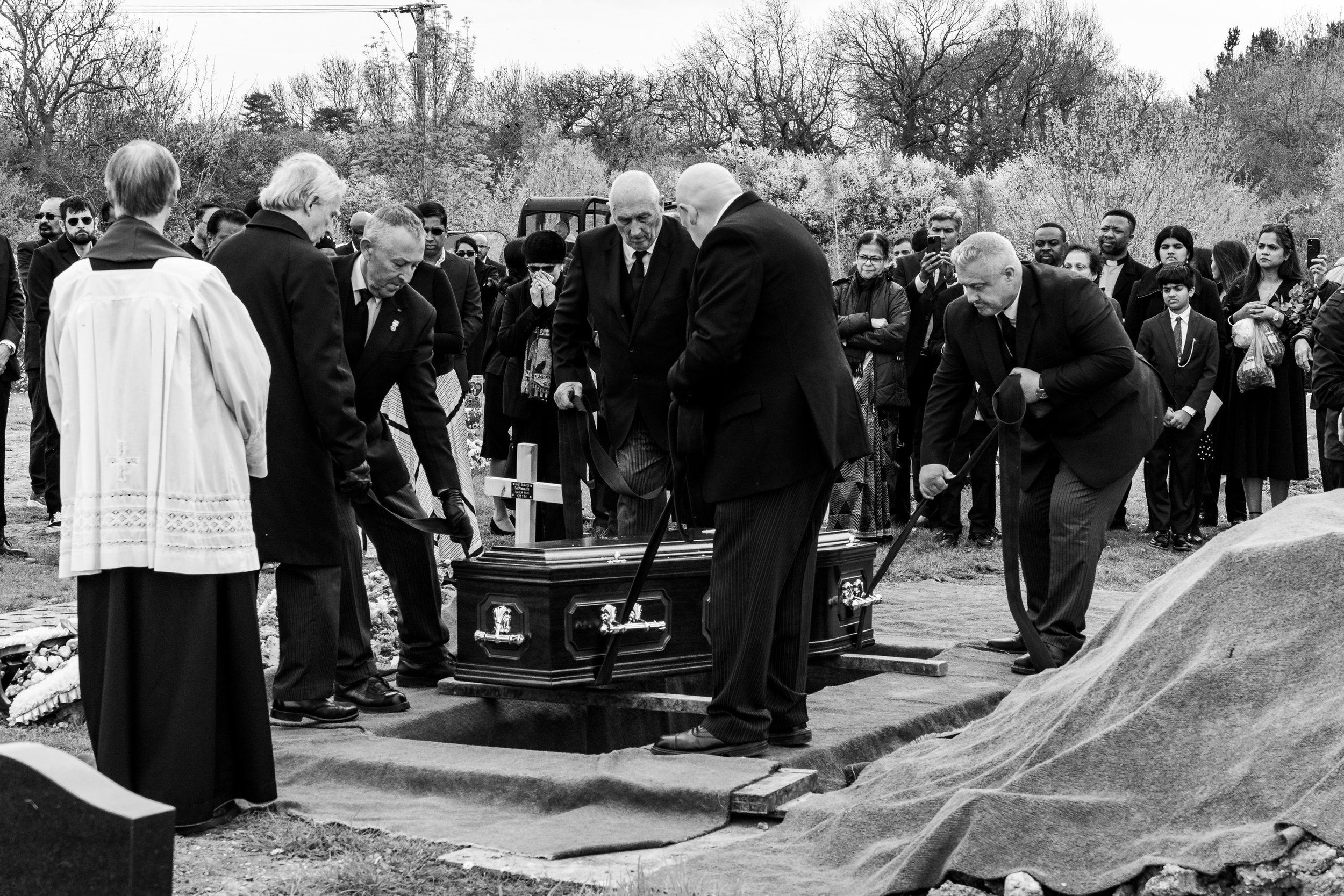 Pallbearers lower a coffin into the ground as an officiant and congregation look on at a graveside funeral service, black and white documentary photography by Paul Hands