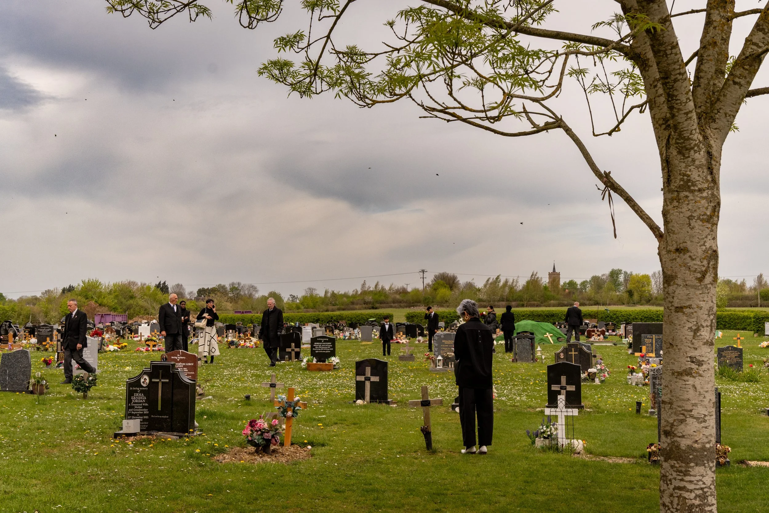 Mourners dressed in black gathered among headstones in a cemetery on an overcast spring day, funeral photography by Paul Hands