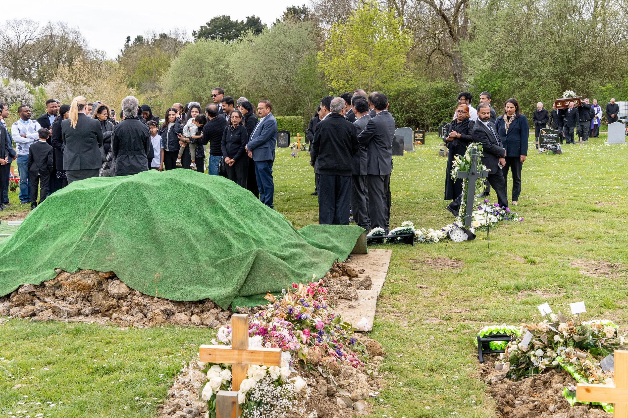 A large multi-cultural congregation gathered around a graveside at an outdoor funeral ceremony, flowers laid on and around the grave, funeral photography by Paul Hands