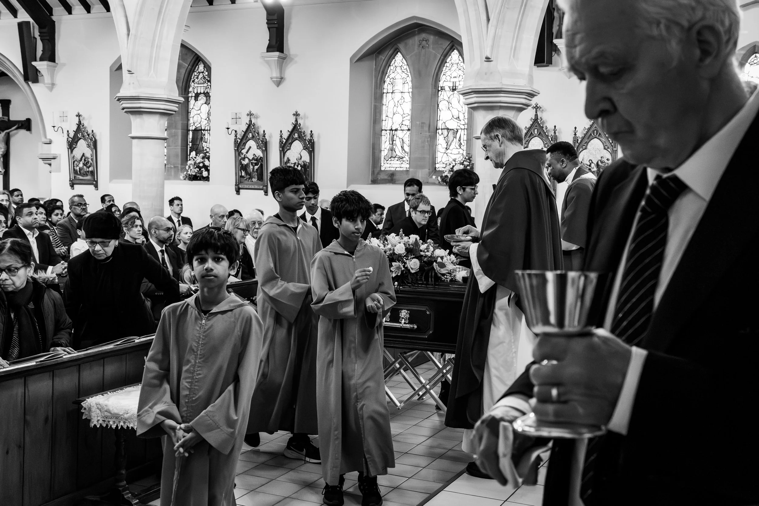 Children in ceremonial robes processing down a church aisle past the coffin during a multi-faith funeral service, black and white documentary photography by Paul Hands