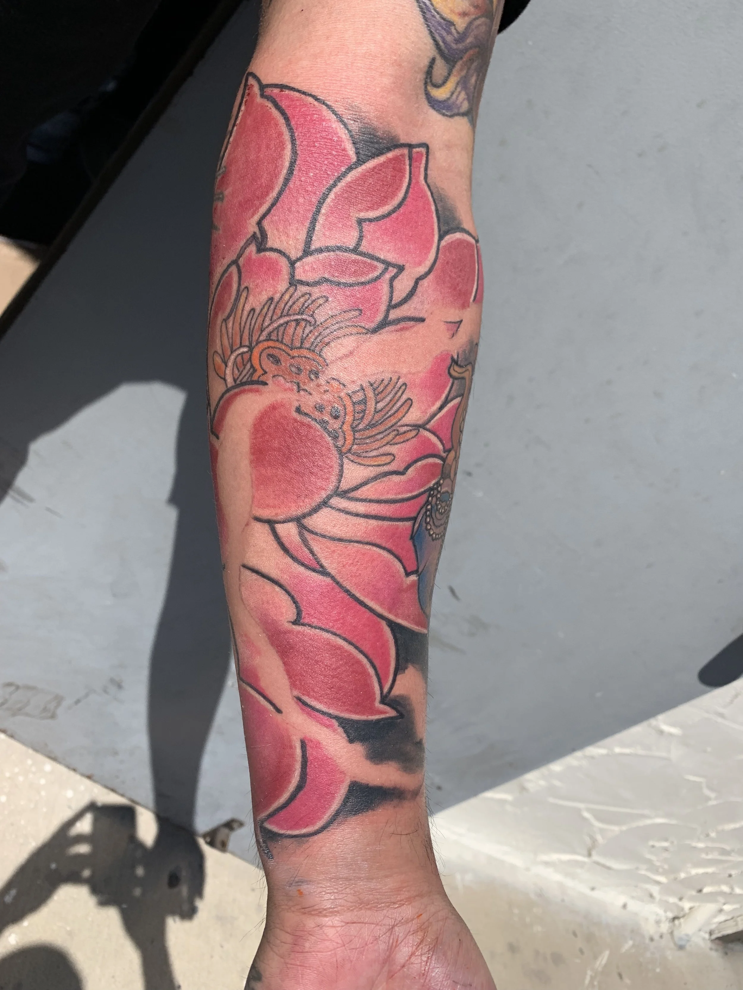 Tattoo of a pink flower with black outline on a person's forearm, with additional tattooed design visible above and to the side.