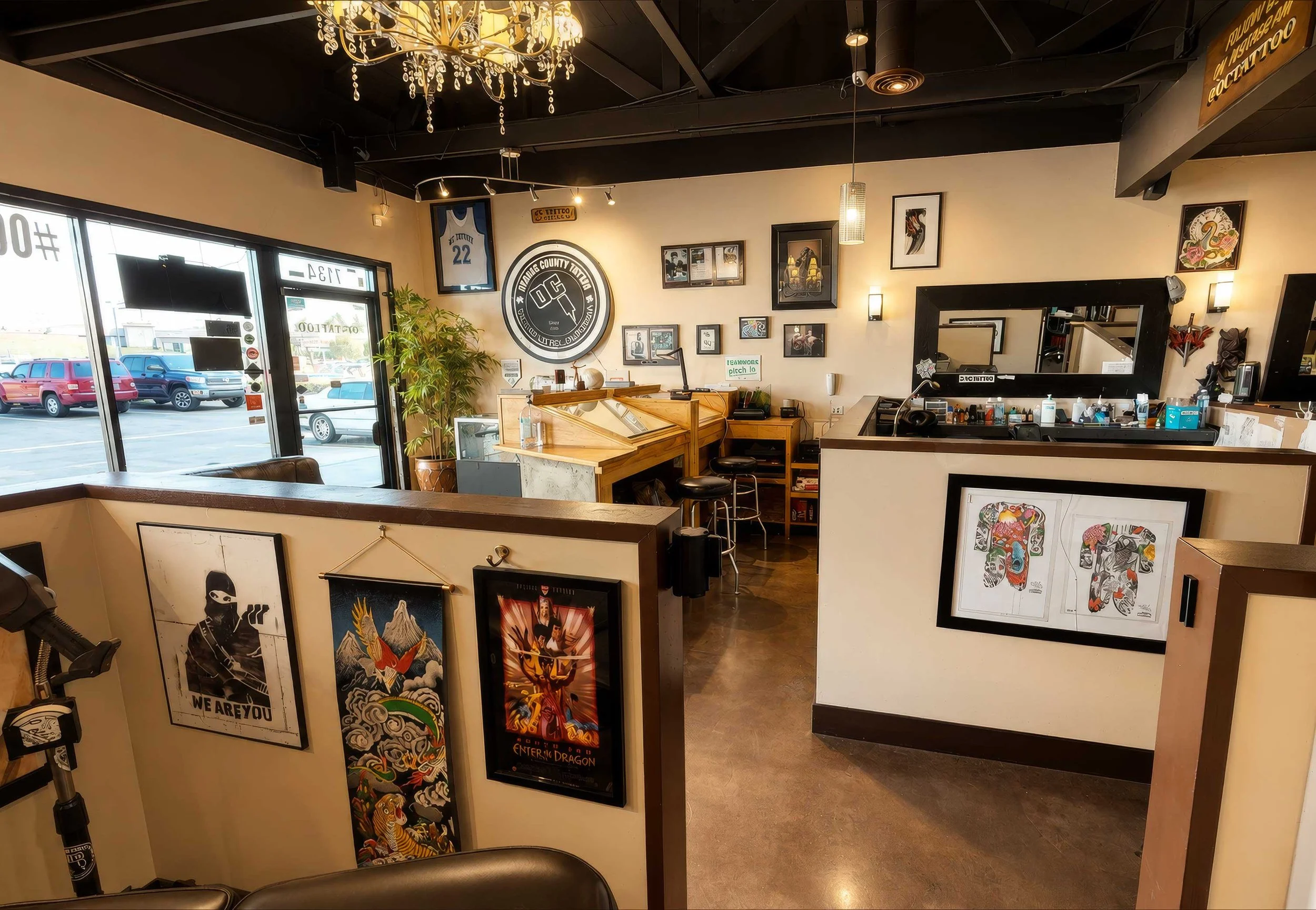 Interior of a tattoo shop with framed artwork on the walls, a large logo on the center wall, a wooden reception counter, and various decor items, including a chandelier and plants, with large windows showing parked cars outside.
