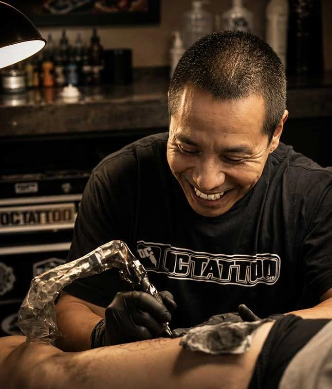A tattoo artist smiling while tattooing a person's arm in a studio.