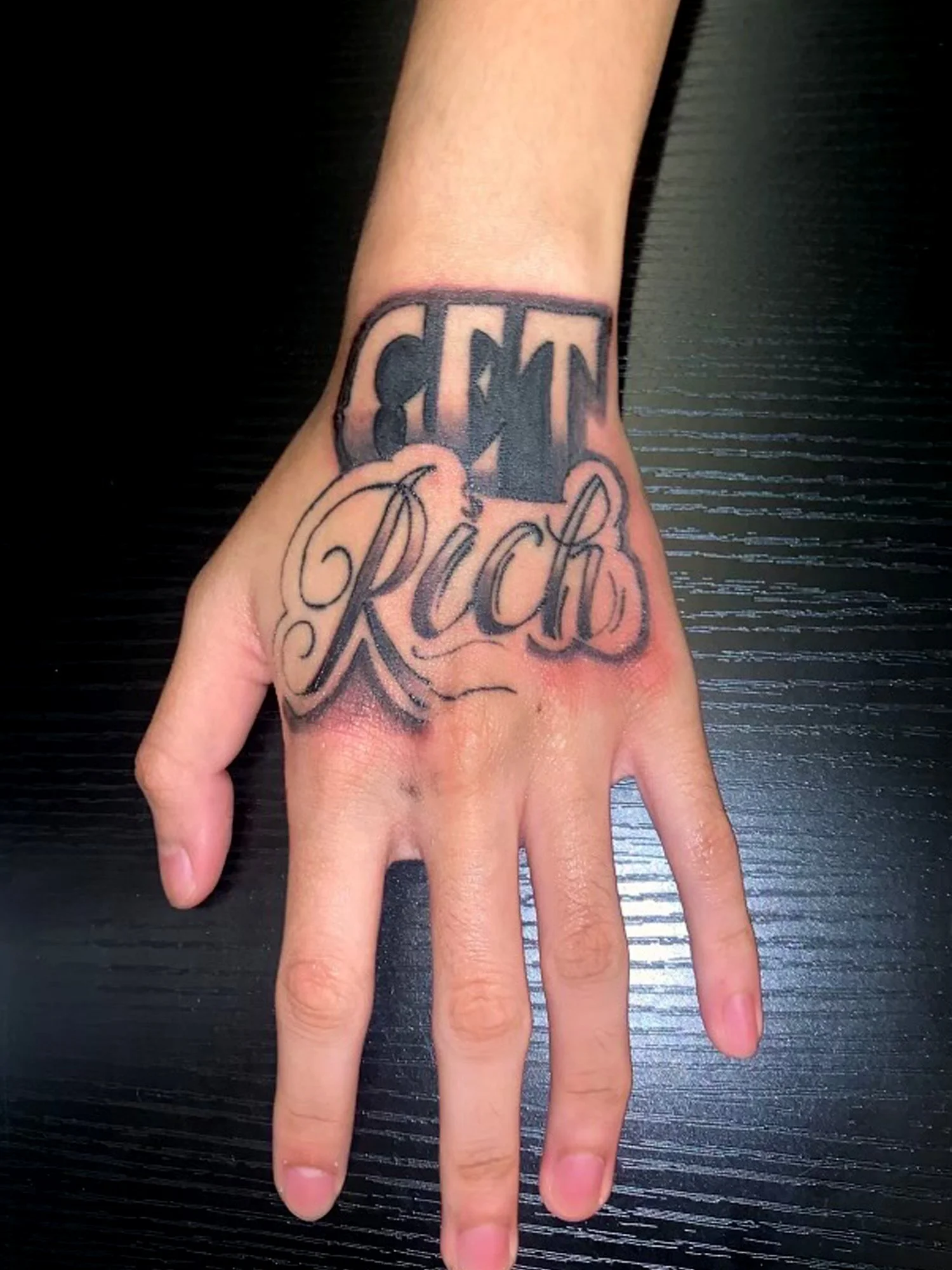 Tattoo on a person's hand with the words 'Chant' in bold letters above 'Rich' in cursive script, on a black and textured surface.