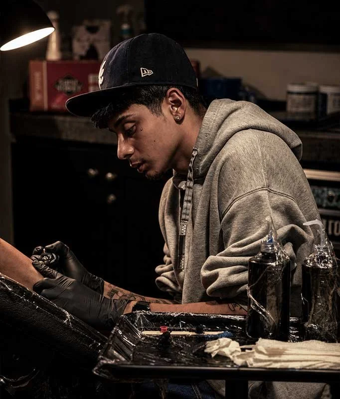 Young man in a gray hoodie and black cap getting a tattoo on his arm in a tattoo parlor.