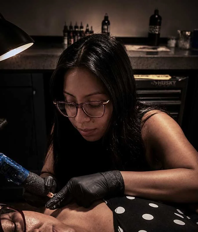 A woman with black hair and glasses, wearing black gloves, is doing a tattoo on a person's upper arm in a dimly lit room with tattoo supplies on the counter behind her.