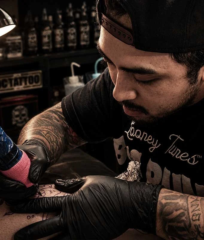 Tattoo artist wearing black gloves and a black cap, tattooing a person's arm in a tattoo shop.