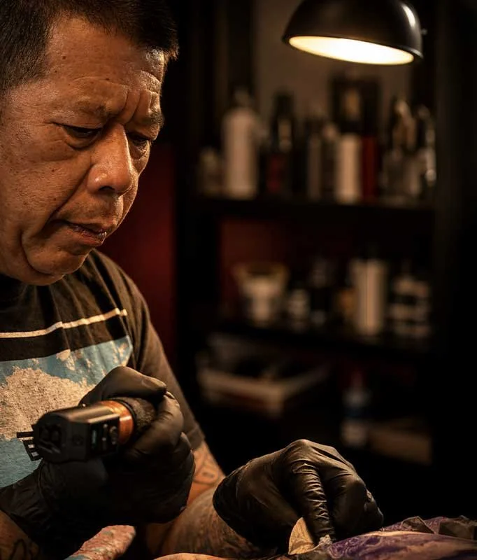 A tattoo artist in a dimly lit room tattooing a person's arm. The artist is wearing black gloves and holding a tattoo machine, focusing on his work.