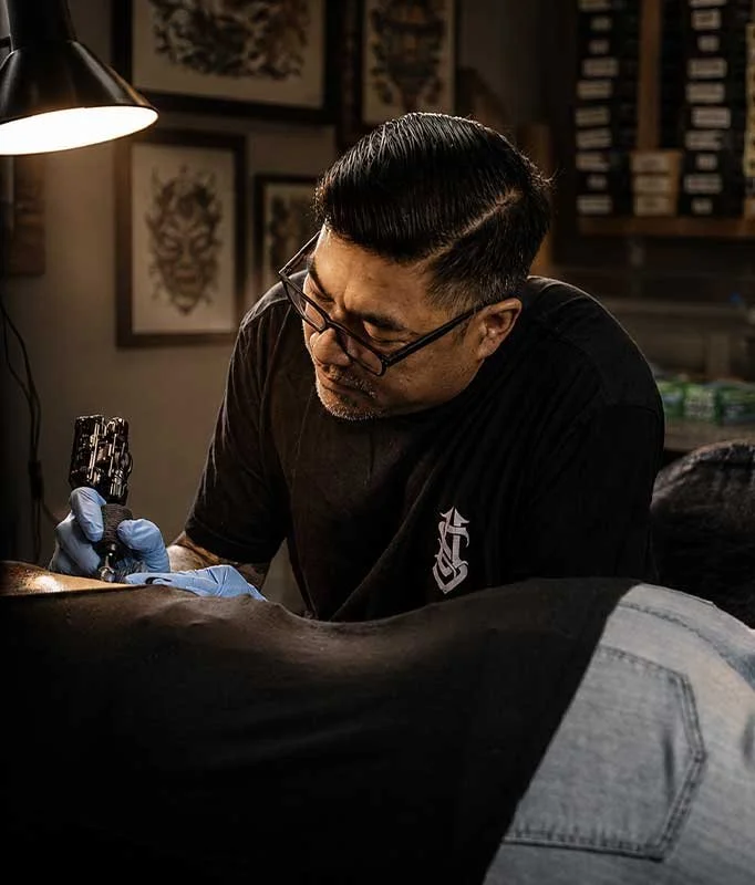 Tattoo artist wearing glasses and gloves working on a client's arm in a tattoo studio, with framed artwork on the wall behind.