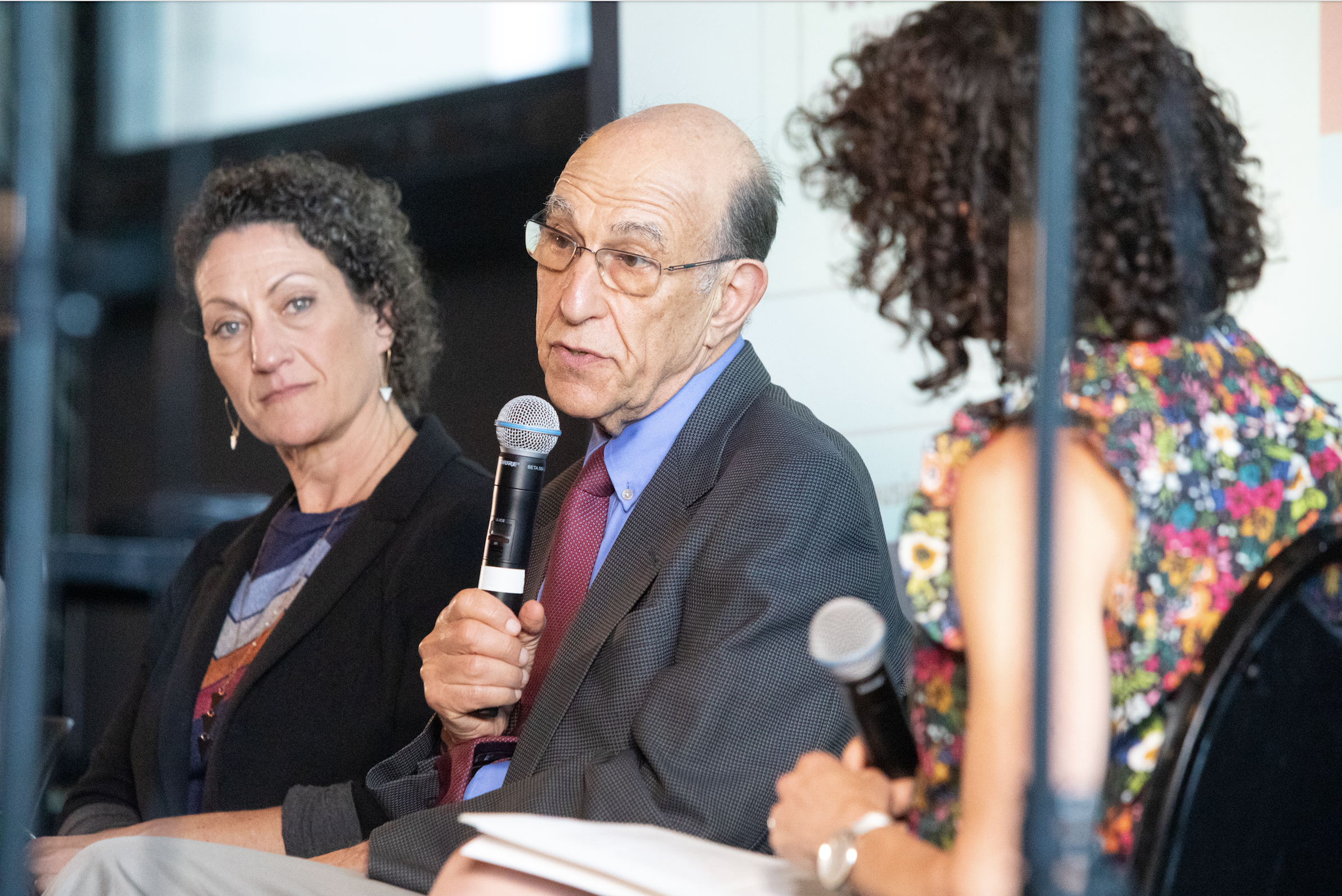  Chicago Housing Commissioner Marisa Novara sits next to Leah and Richard Rothstein. Richard Rothstein speaks into a microphone.  