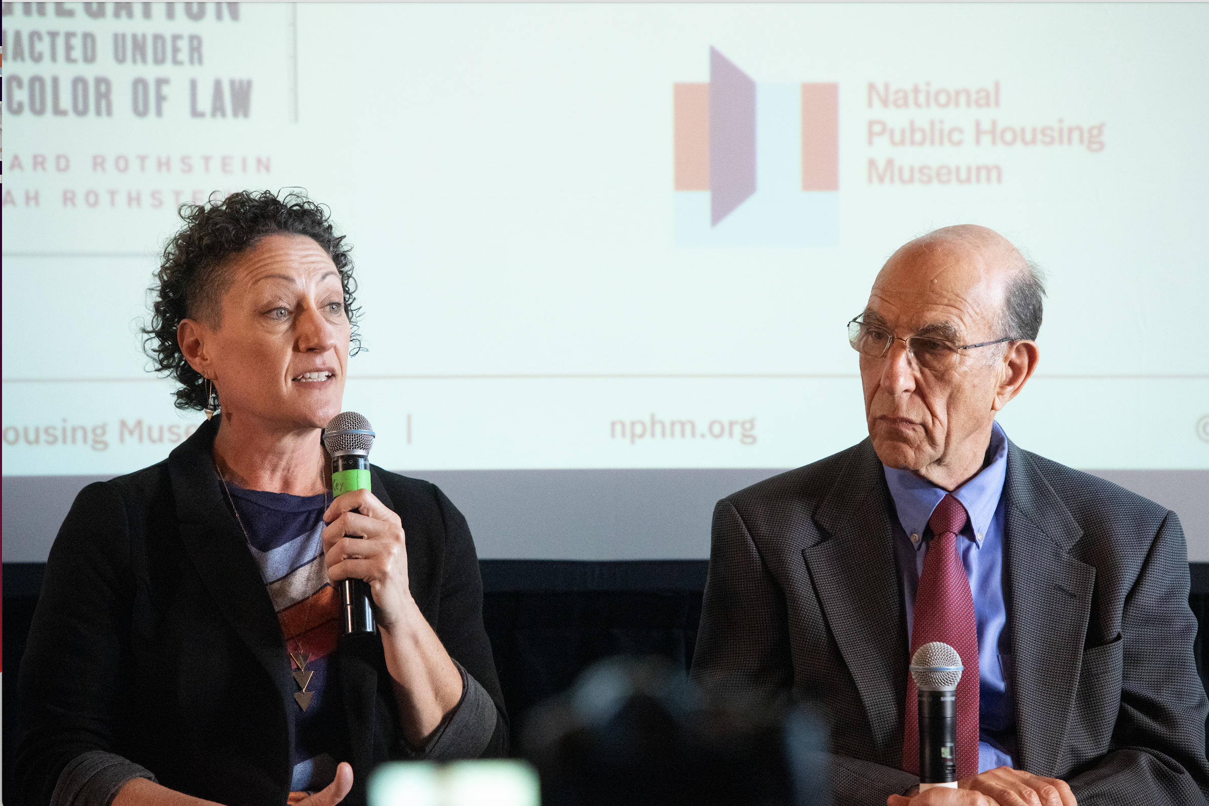  Leah Rothstein speaks into a microphone while sitting in front of a screen with the National Public Housing Museum’s logo on it. Richard Rothstein sits next to her. 