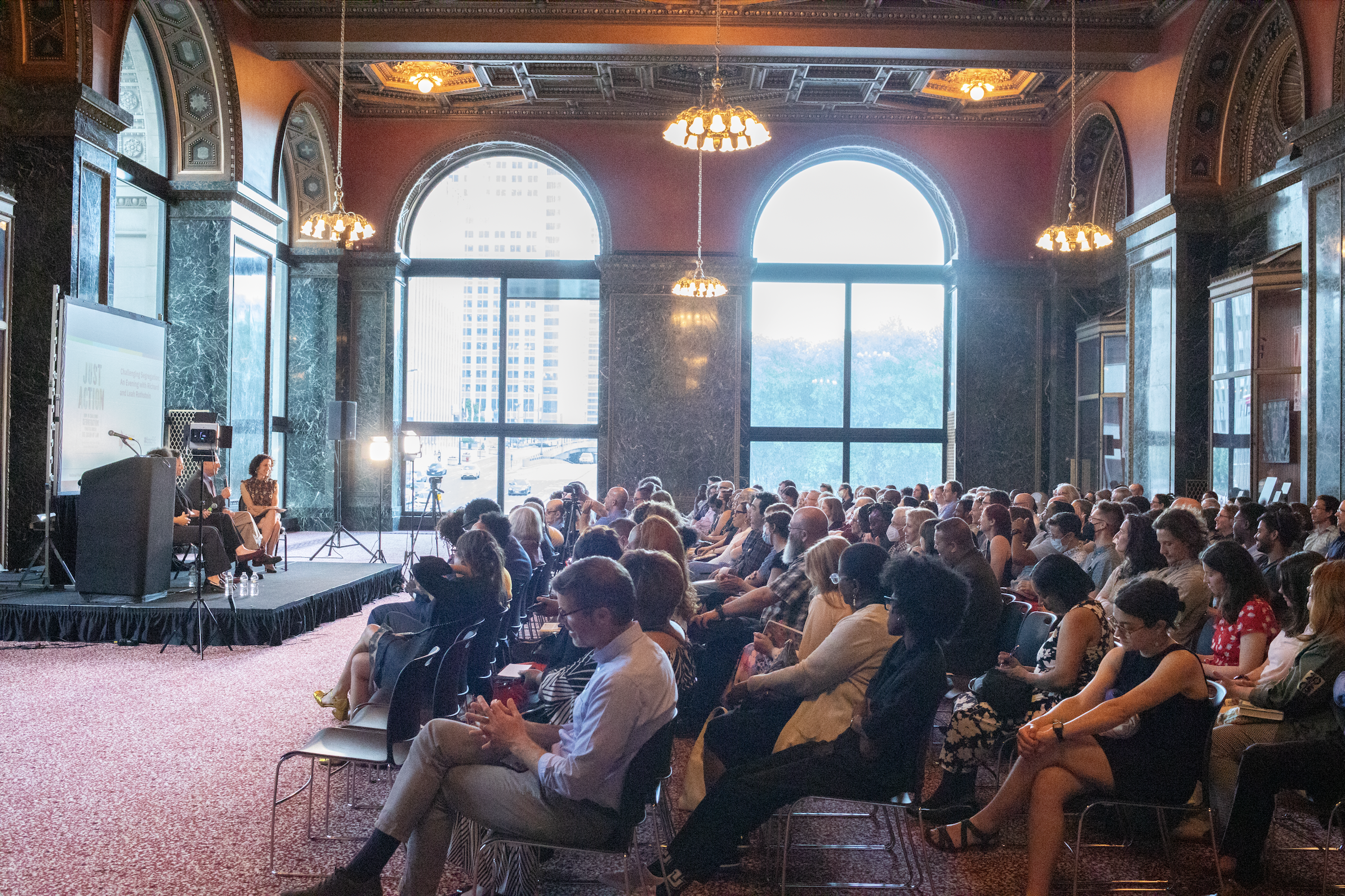  A crowd sits in the Chicago Cultural Center’s G.A.R. Hall, an ornate room with soaring ceilings and large windows. 