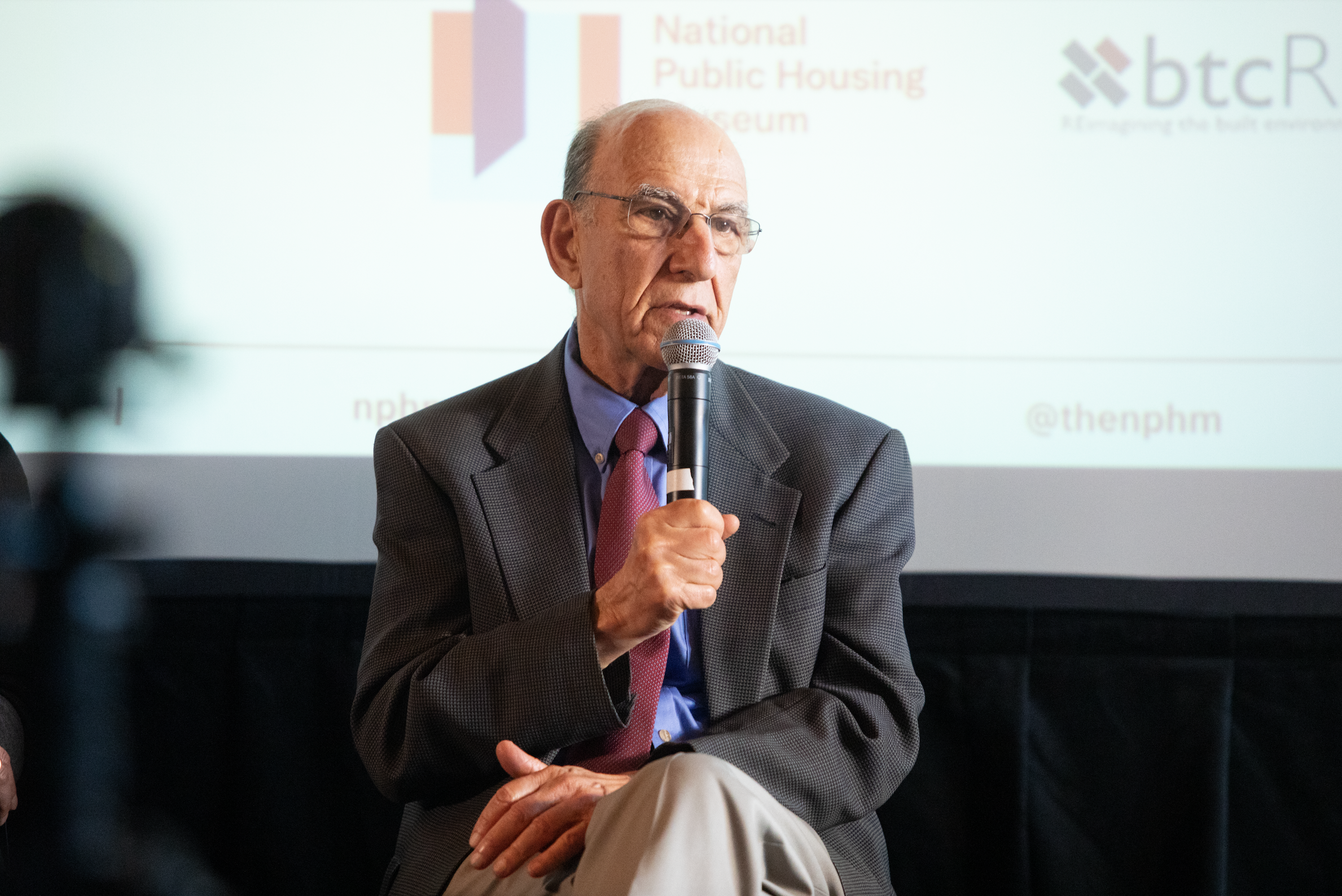  Richard Rothstein speaks into a microphone while seated.  